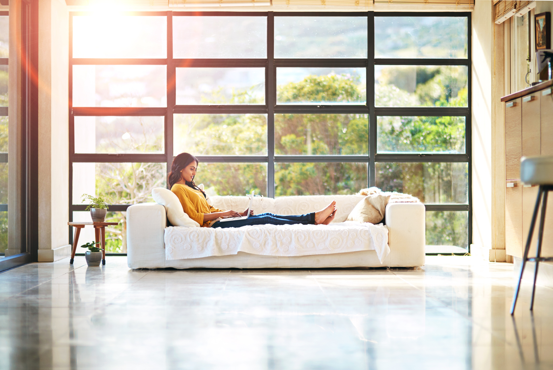 Woman relaxing on a sofa reading on her laptop in a bright, sunlit room